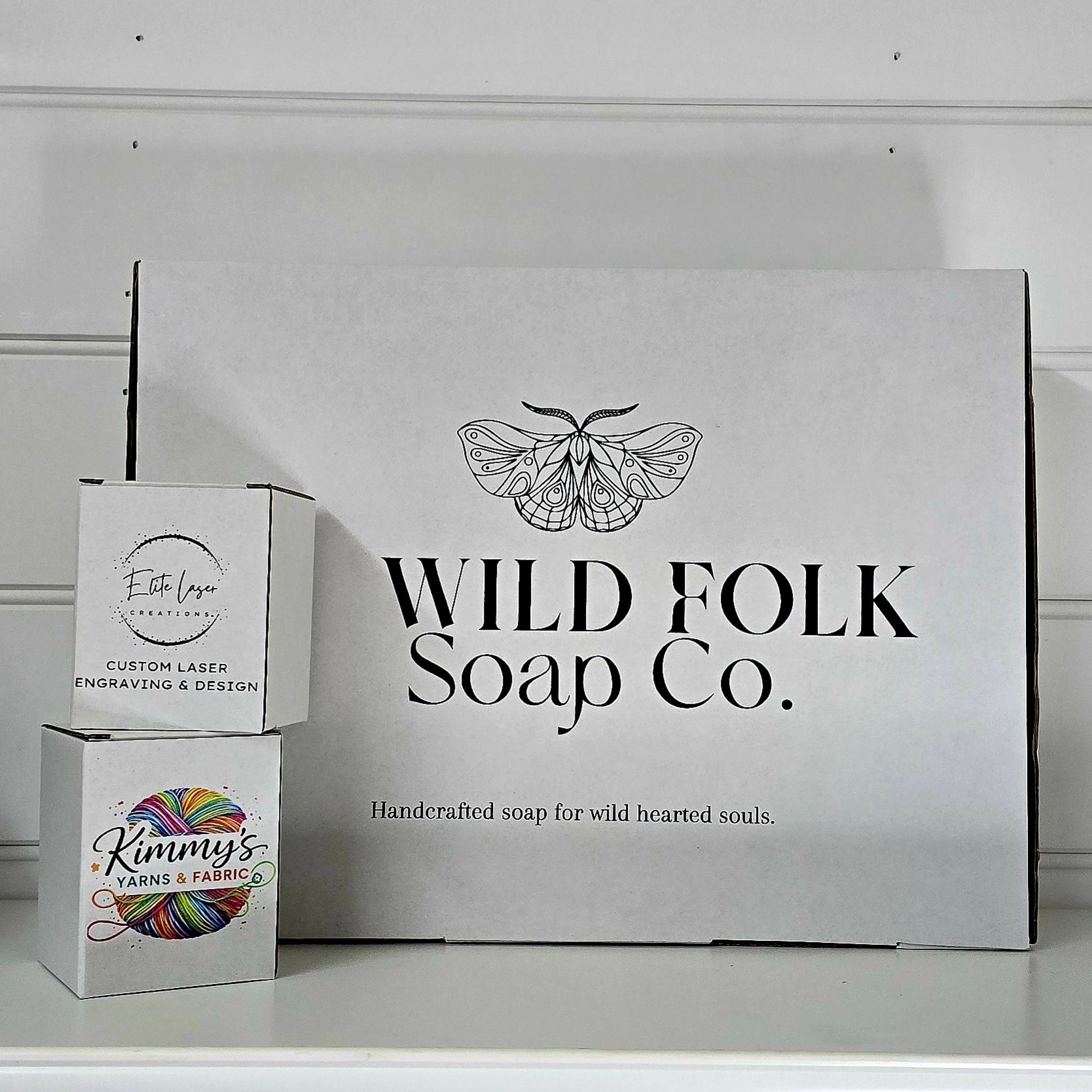 A large white cardboard box for Wild Folk Soap Co. with a black moth logo and text, standing on a white surface next to two smaller branded boxes stacked on top of each other.
