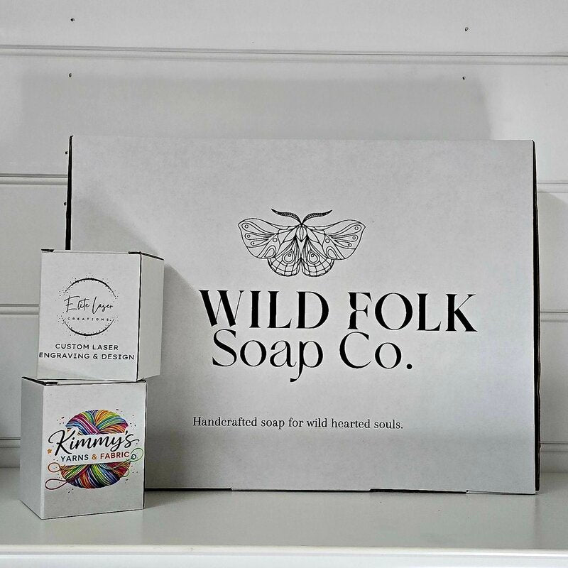 A large white cardboard box for Wild Folk Soap Co. with a black moth logo and text, standing on a white surface next to two smaller branded boxes stacked on top of each other.