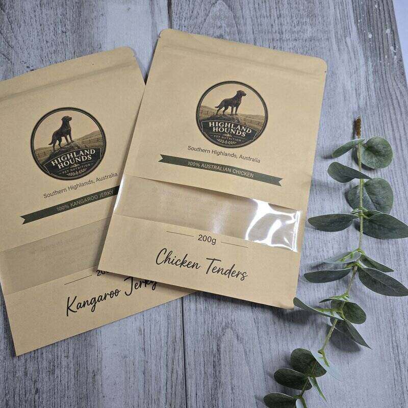 Two brown kraft paper pouches of Highland Bounds Chicken Tenders dog food, displayed on a gray wood-textured surface with a green eucalyptus sprig.