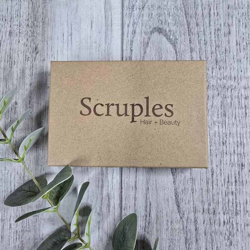 A square kraft paper box labeled 'Scruples Hair + Beauty' in dark brown lettering, placed on a light gray wood plank surface with green leaves at the bottom left.