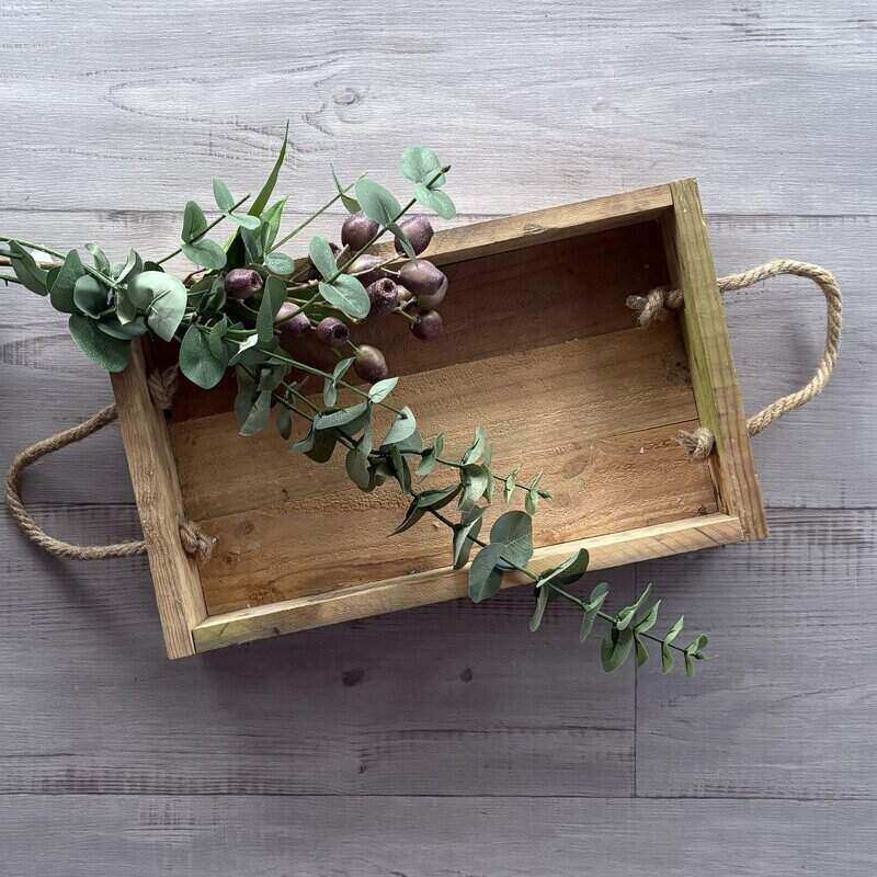 A handmade wooden box with twisted rope handles and a rustic look, placed on a grey surface with some greenery placed on top.