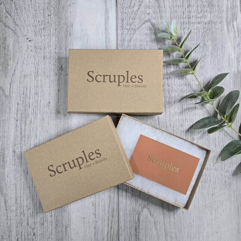 Three Scruples Hair + Beauty branded kraft paper boxes arranged on a light gray wood surface, with one open box showing a coral-orange card and greenery sprig nearby.