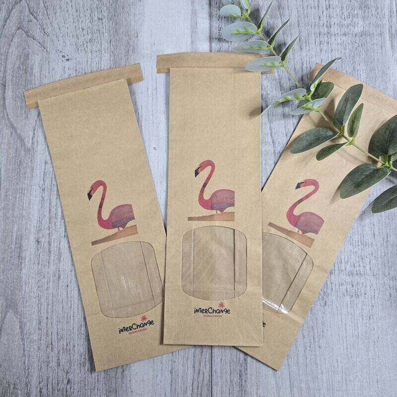 Three kraft paper gift bags with clear window cutouts and a pink flamingo illustration, arranged on a gray wood-textured surface with green leaves.