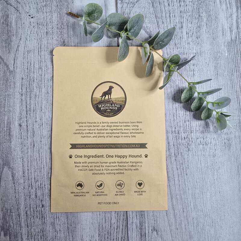 A flat-lay photograph of a Highland Hounds pet food pouch on a gray wood-textured background, decorated with green eucalyptus branches. The pouch is a light tan kraft paper color with black text and a circular logo at the top center.