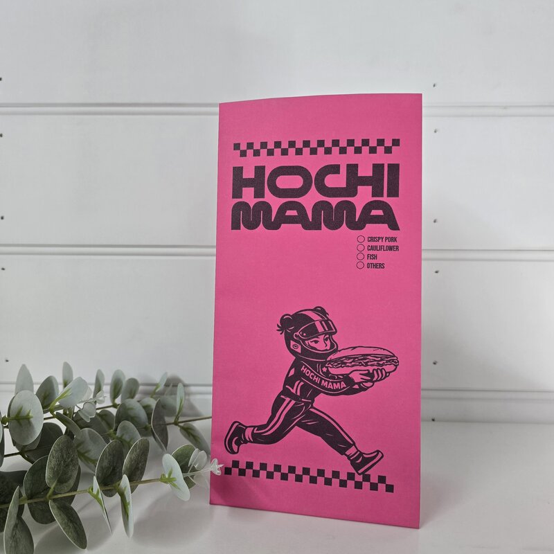 A bright pink novelty coffee bag labeled 'HOCHI MAMA' with a black graphic of a race car driver holding a pizza, standing on a white surface against a white paneled wall with green leaves at the side.