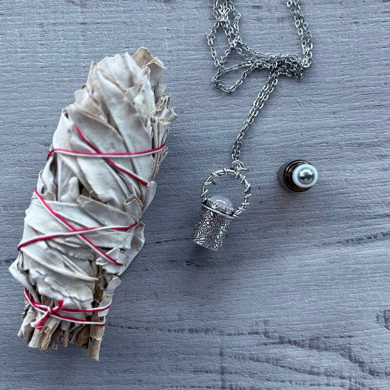 Amber glass roller with ornate silver lid with rose quartz crystal stone & silver chain necklace next to roller pictured with sage smudge stick