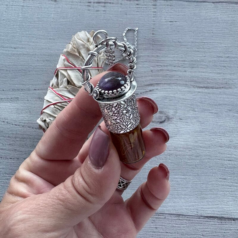 Amber glass roller with ornate silver lid with amethyst crystal stone & silver chain necklace pictured held in hand with sage smudge stick in background