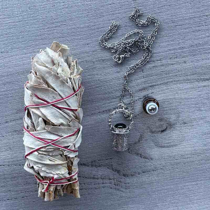 Amber glass roller with ornate silver lid with black onyx crystal stone & silver chain necklace next to roller pictured with sage smudge stick