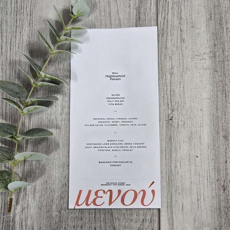 A minimalist white paper menu card with black text and a red 'μενού' at the bottom, photographed on a weathered gray wood surface with green eucalyptus leaves along the left side.